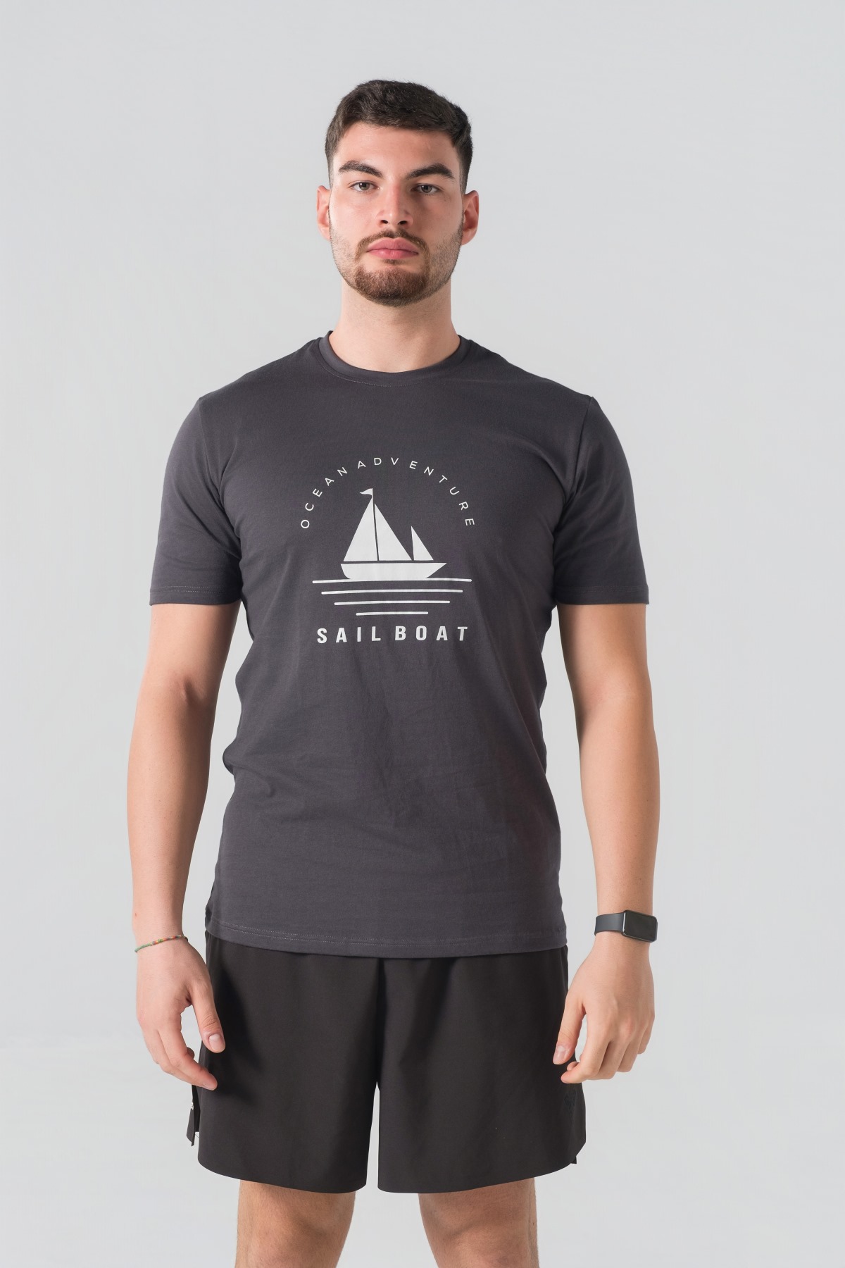 T-shirt Breezy Unisex – Bluzë e thjeshtë me mëngë të shkurtra unisex në ngjyrë hiri të mbyllur (dark grey/charcoal), me jakë të rrumbullakët, print grafik veror detar me anije me vela dhe mbishkrimin "Ocean Adventure / Sail Boat" në ngjyrë të bardhë në gjoks.