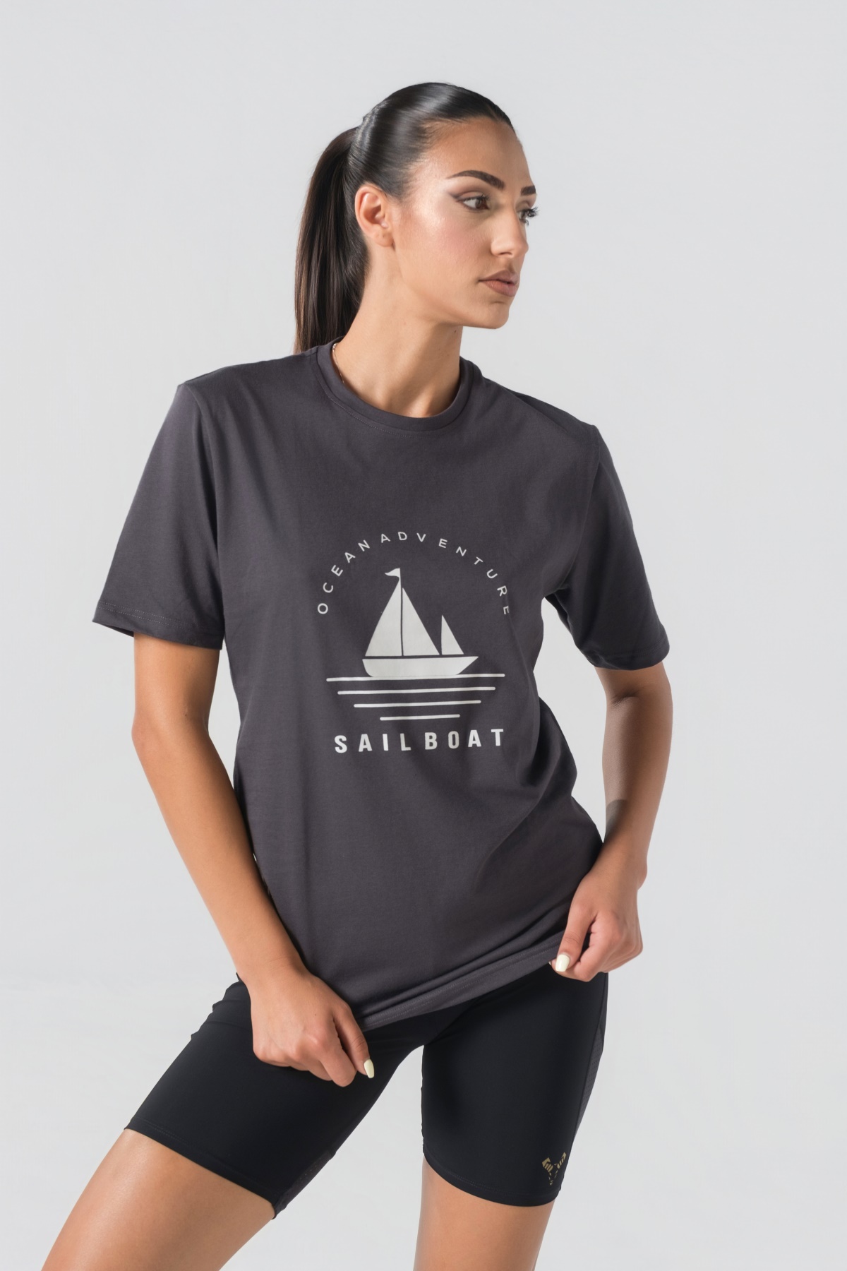 T-shirt Breezy Unisex – Bluzë e thjeshtë me mëngë të shkurtra unisex në ngjyrë hiri të mbyllur (dark grey/charcoal), me jakë të rrumbullakët, print grafik veror detar me anije me vela dhe mbishkrimin "Ocean Adventure / Sail Boat" në ngjyrë të bardhë në gjoks.
