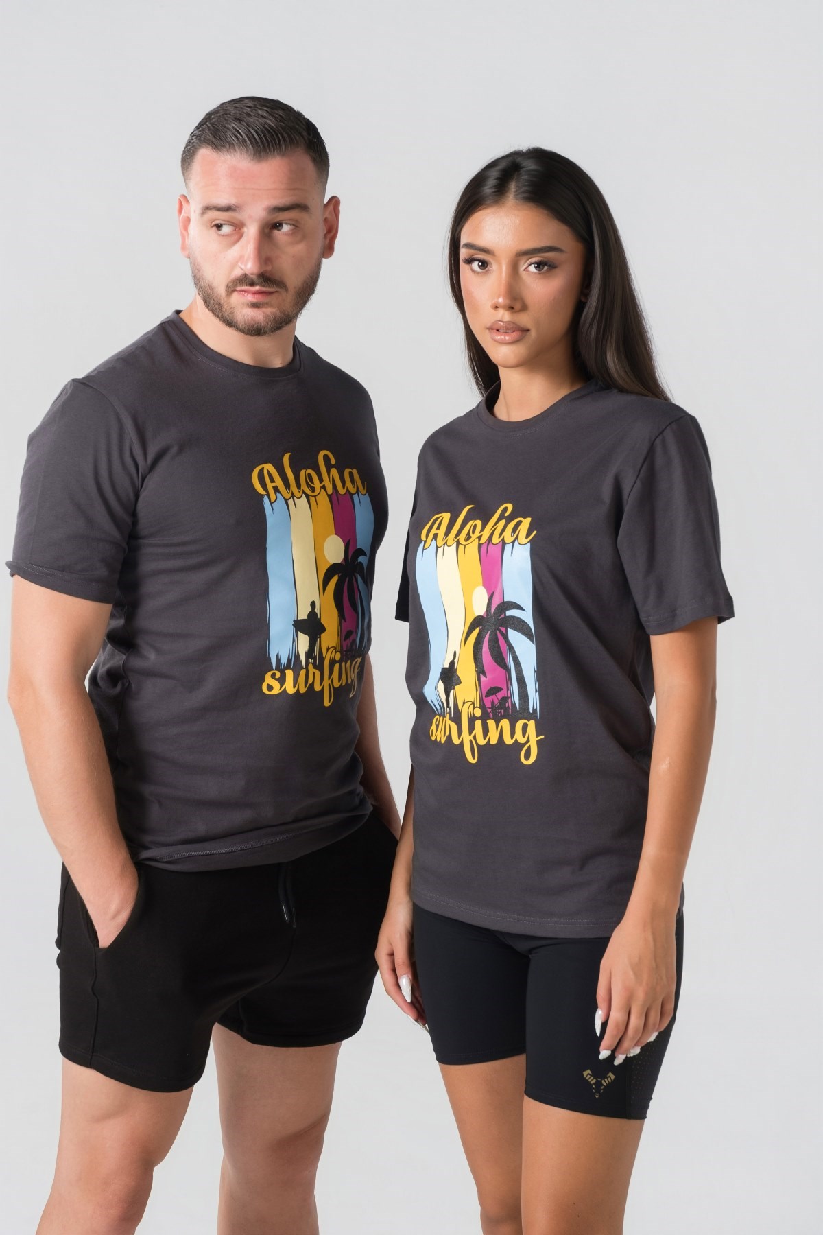 T-shirt Breezy Unisex – Bluzë e thjeshtë me mëngë të shkurtra unisex në ngjyrë hiri të mbyllur (dark grey/charcoal), me jakë të rrumbullakët, print grafik veror tropikal me palma, surfist dhe mbishkrimin "Aloha Surfing" në ngjyra të gjalla në gjoks.