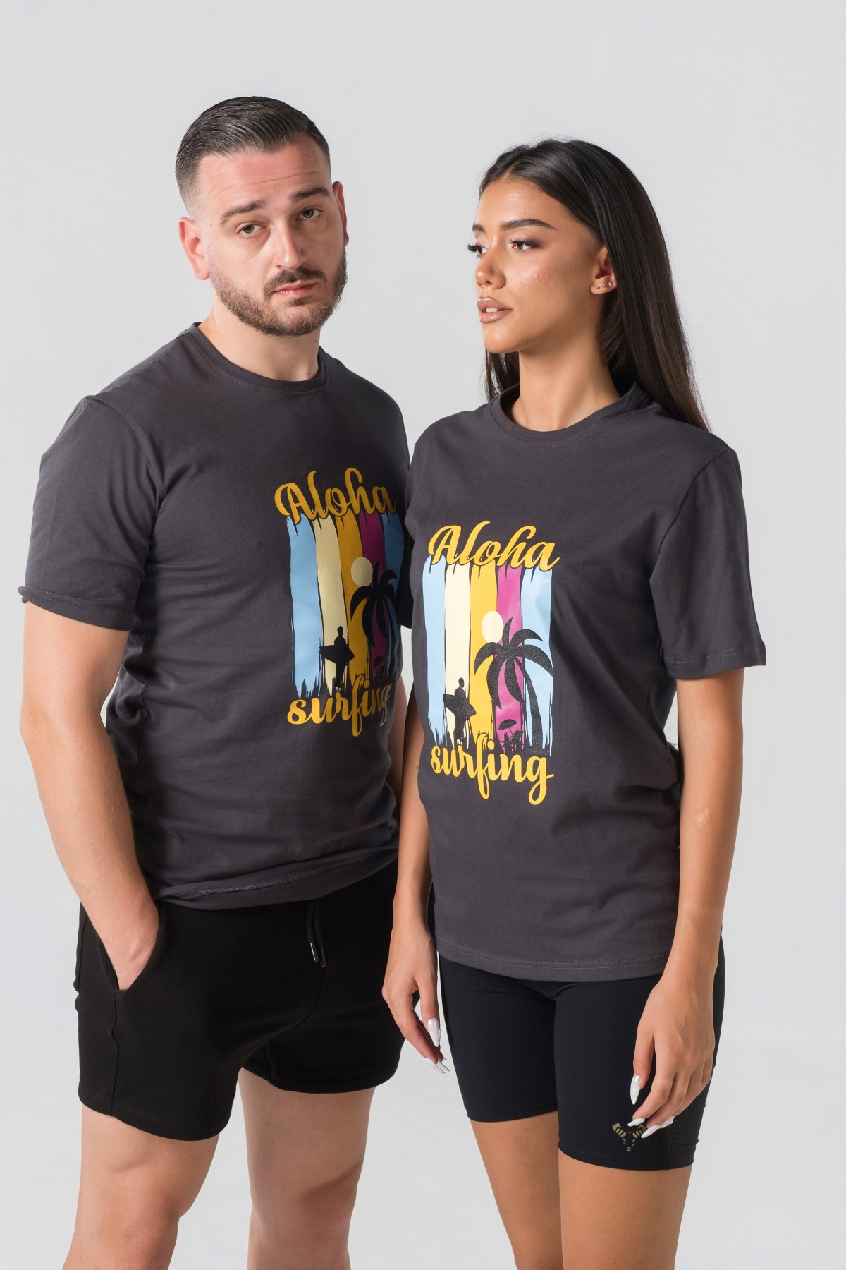 T-shirt Breezy Unisex – Bluzë e thjeshtë me mëngë të shkurtra unisex në ngjyrë hiri të mbyllur (dark grey/charcoal), me jakë të rrumbullakët, print grafik veror tropikal me palma, surfist dhe mbishkrimin "Aloha Surfing" në ngjyra të gjalla në gjoks.