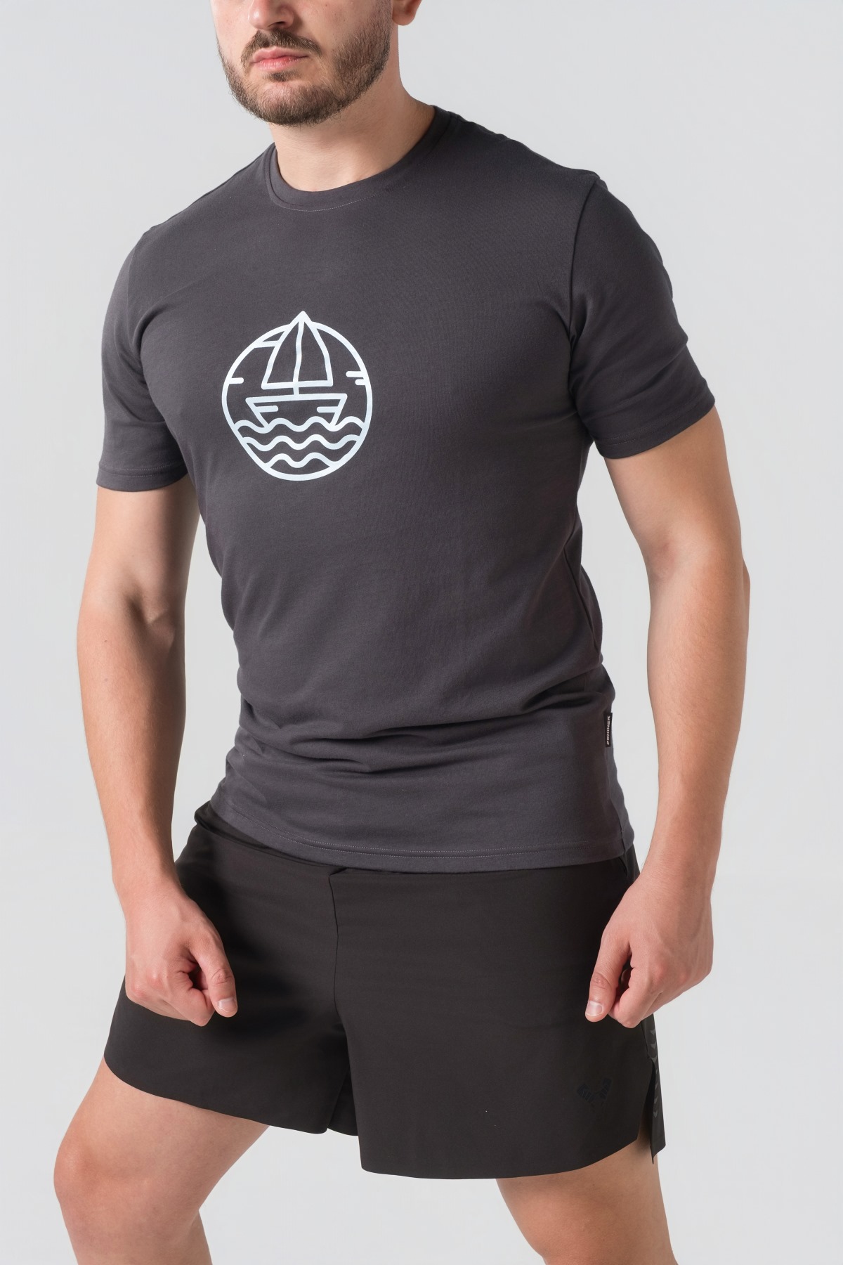 T-shirt Breezy Unisex – Bluzë e thjeshtë me mëngë të shkurtra unisex në ngjyrë hiri të mbyllur (dark grey/charcoal), me jakë të rrumbullakët, print grafik me emblemë rrethore detare me anije/varkë dhe valë deti në ngjyrë të kaltër të çelët në gjoks.