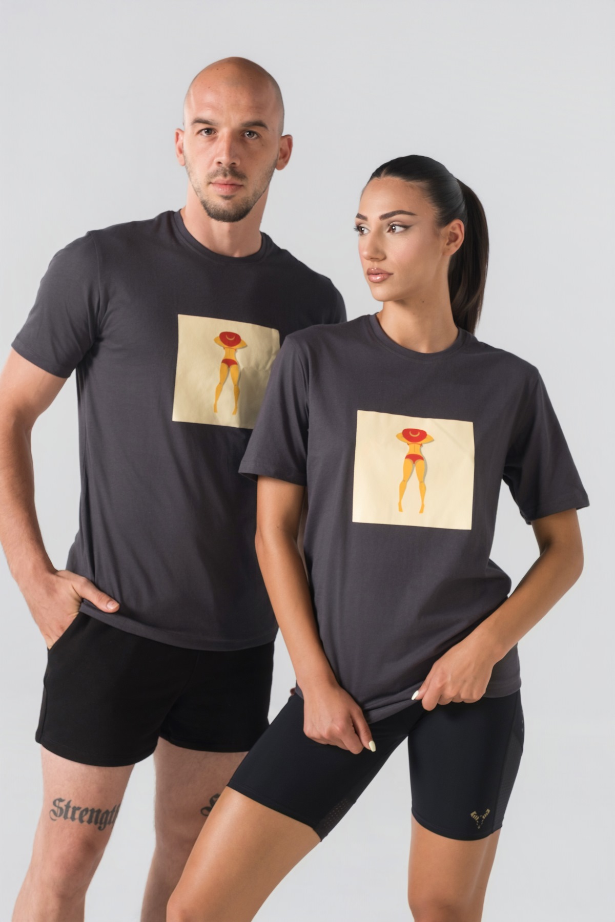 T-shirt Breezy Unisex – Bluzë e thjeshtë me mëngë të shkurtra unisex në ngjyrë hiri të mbyllur (dark grey/charcoal), me jakë të rrumbullakët, print grafik katror veror me një femër duke u rrezitur në plazh me bikini të kuqe dhe kapelë të kuqe në gjoks.