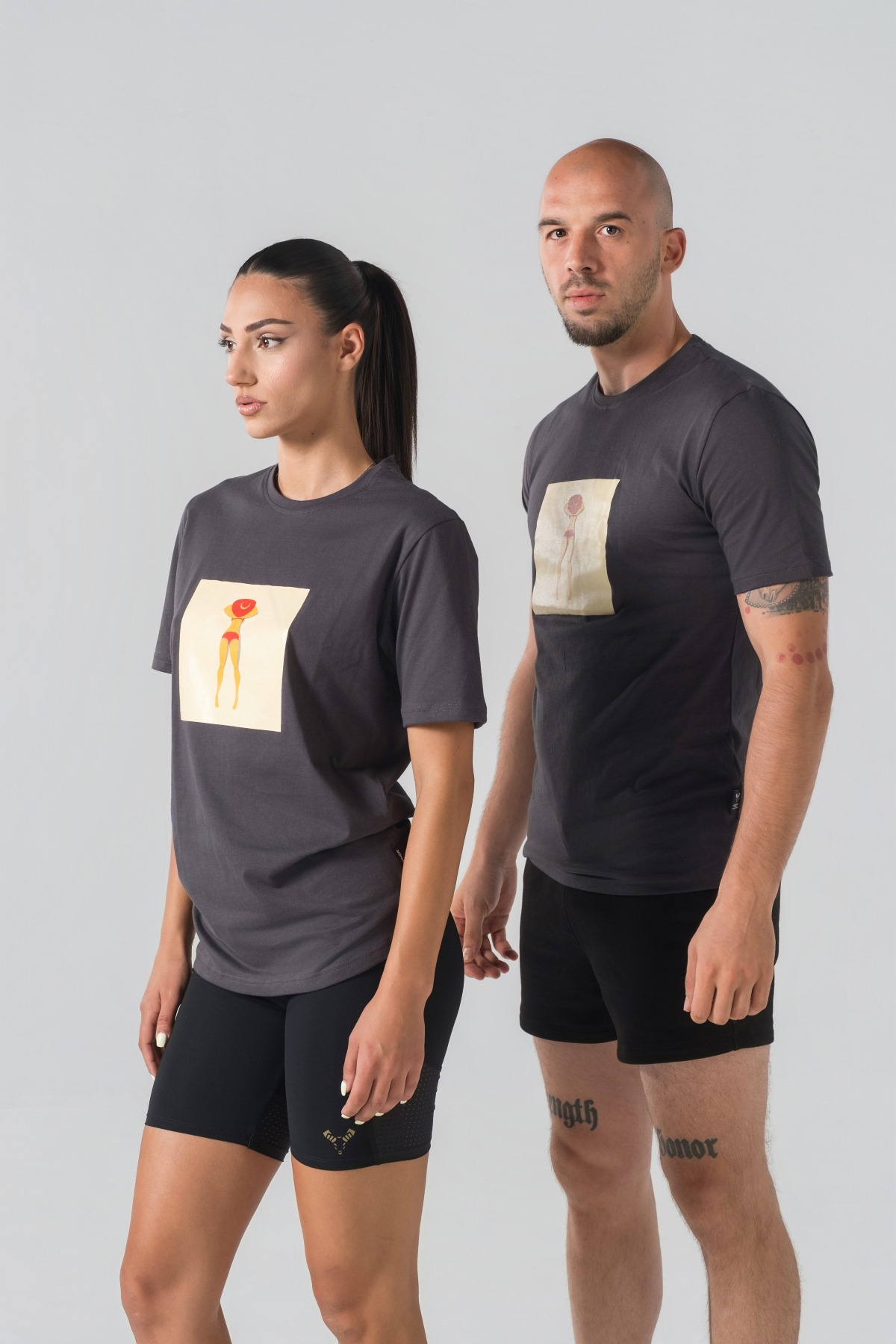 T-shirt Breezy Unisex – Bluzë e thjeshtë me mëngë të shkurtra unisex në ngjyrë hiri të mbyllur (dark grey/charcoal), me jakë të rrumbullakët, print grafik katror veror me një femër duke u rrezitur në plazh me bikini të kuqe dhe kapelë të kuqe në gjoks.