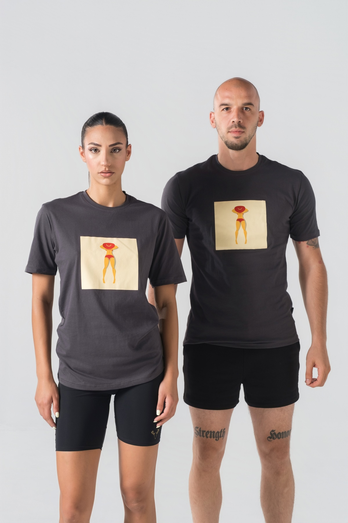 T-shirt Breezy Unisex – Bluzë e thjeshtë me mëngë të shkurtra unisex në ngjyrë hiri të mbyllur (dark grey/charcoal), me jakë të rrumbullakët, print grafik katror veror me një femër duke u rrezitur në plazh me bikini të kuqe dhe kapelë të kuqe në gjoks.
