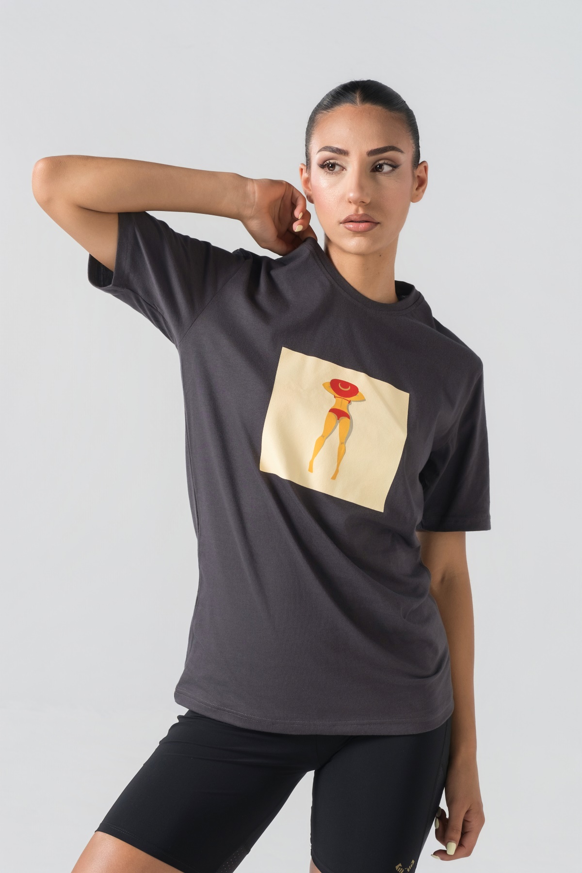 T-shirt Breezy Unisex – Bluzë e thjeshtë me mëngë të shkurtra unisex në ngjyrë hiri të mbyllur (dark grey/charcoal), me jakë të rrumbullakët, print grafik katror veror me një femër duke u rrezitur në plazh me bikini të kuqe dhe kapelë të kuqe në gjoks.