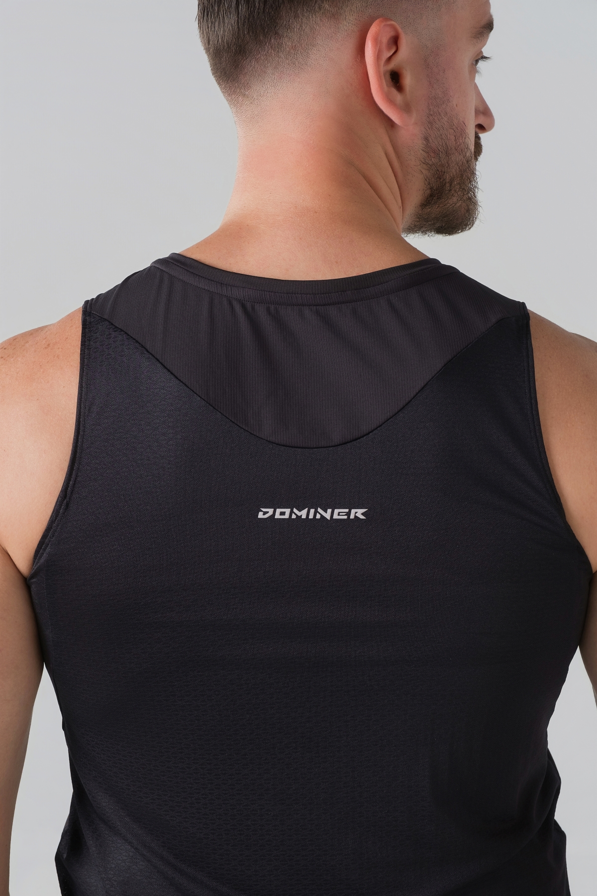 Shift Kanatjere Meshkujsh – Kanatjere sportive për meshkuj në ngjyrë të zezë, me jakë të rrumbullakët, material me teksturë rrjetë (mesh) në shpinë për ajrosje, të çara të vogla anësore në fund, logo minimale Dominer në gjoks, emrin e markës të printuar në pjesën e pasme sipër dhe detaj vertikal me shigjeta në pjesën e poshtme prapa.