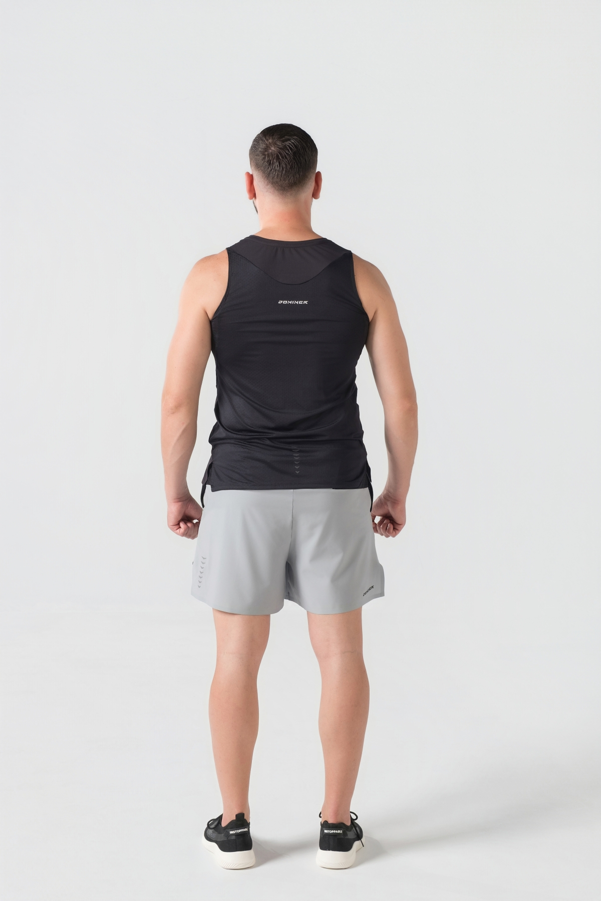 Shift Kanatjere Meshkujsh – Kanatjere sportive për meshkuj në ngjyrë të zezë, me jakë të rrumbullakët, material me teksturë rrjetë (mesh) në shpinë për ajrosje, të çara të vogla anësore në fund, logo minimale Dominer në gjoks, emrin e markës të printuar në pjesën e pasme sipër dhe detaj vertikal me shigjeta në pjesën e poshtme prapa.