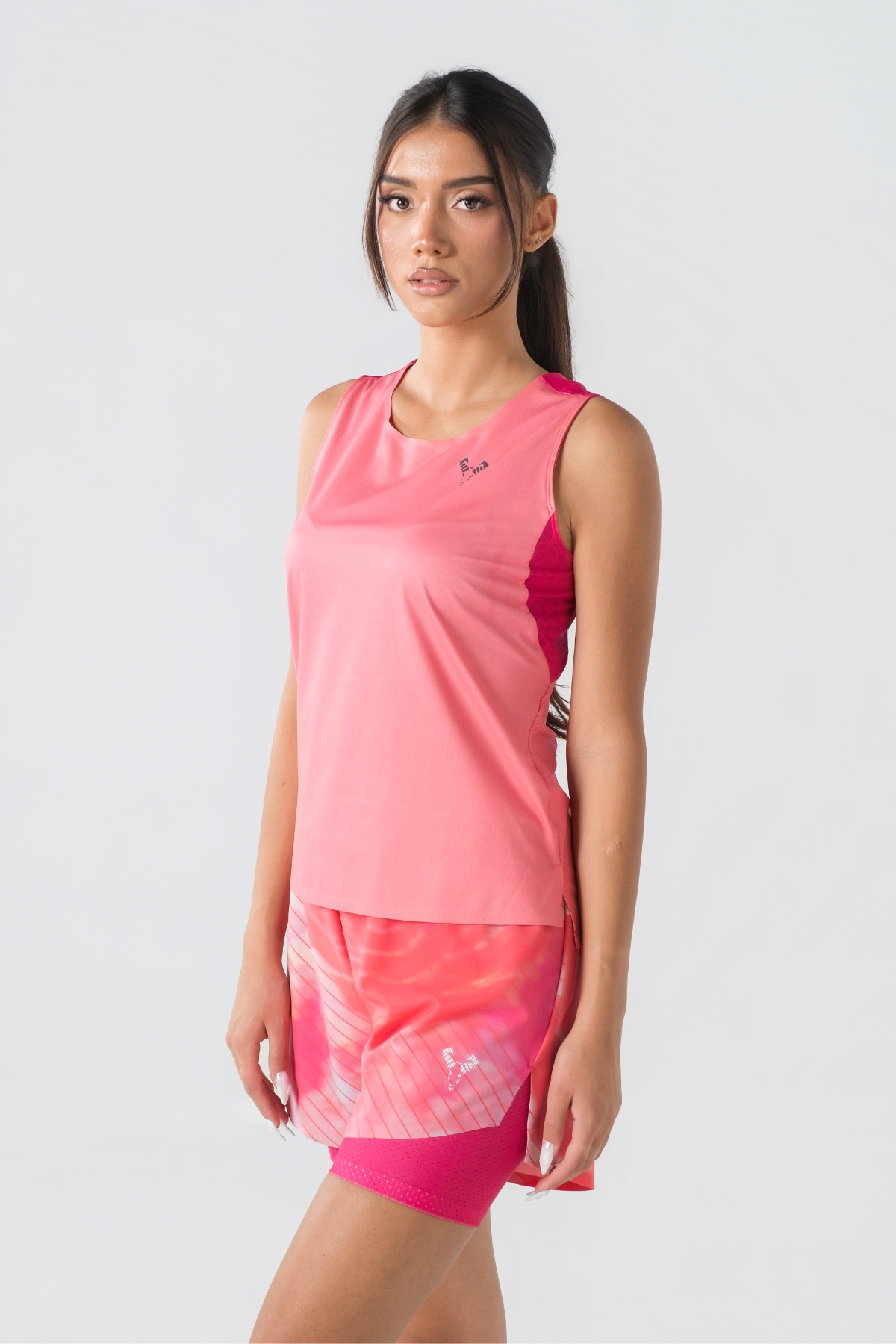 Shift Kanatjere Femrash – Kanatjere sportive për femra në ngjyrë rozë të çelët (coral pink) me panela anësore dhe në shpatulla në ngjyrë rozë më të mbyllur (fuchsia), jakë të rrumbullakët, panel rrjetë (mesh) të shpuar në shpinë për ajrosje, të çara të vogla anësore në fund, logo minimale Dominer në gjoks dhe emrin e markës të printuar në pjesën e pasme sipër.