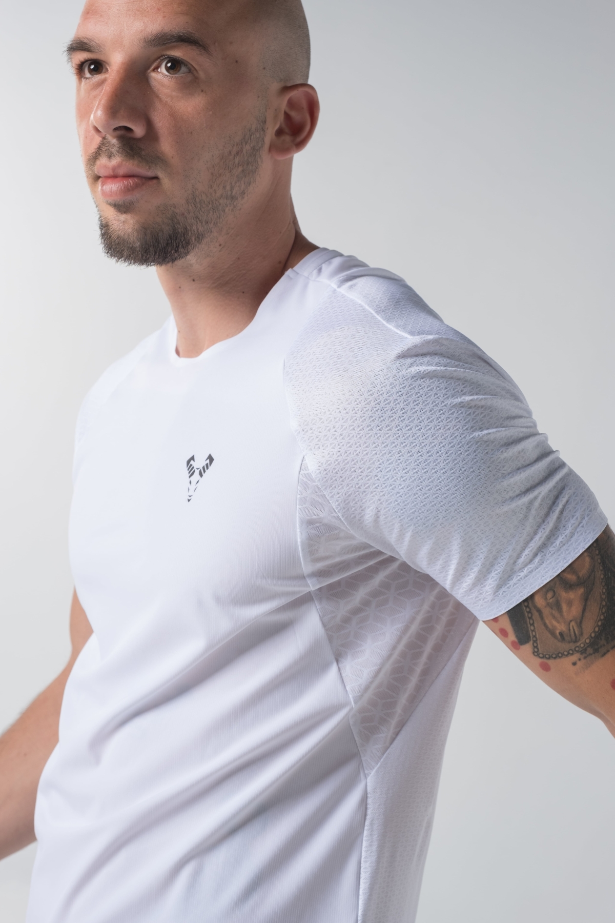 Shift Bluze Meshkujsh – Bluzë sportive me mëngë të shkurtra për meshkuj në ngjyrë të bardhë, me jakë të rrumbullakët, material me teksturë rrjetë për ajrosje, logo minimale Dominer në gjoks, emrin e markës të printuar në pjesën e pasme sipër dhe detaj vertikal me shigjeta në pjesën e poshtme prapa.