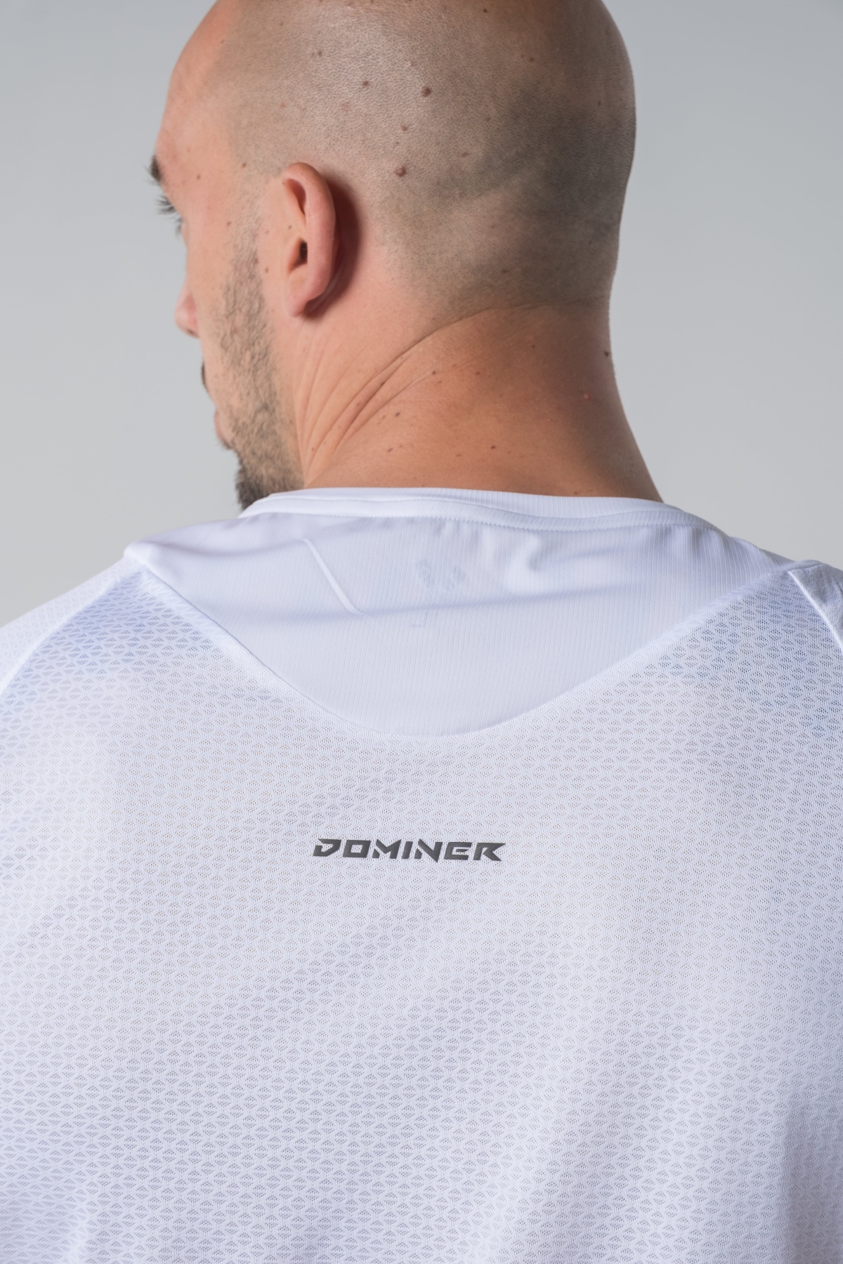 Shift Bluze Meshkujsh – Bluzë sportive me mëngë të shkurtra për meshkuj në ngjyrë të bardhë, me jakë të rrumbullakët, material me teksturë rrjetë për ajrosje, logo minimale Dominer në gjoks, emrin e markës të printuar në pjesën e pasme sipër dhe detaj vertikal me shigjeta në pjesën e poshtme prapa.