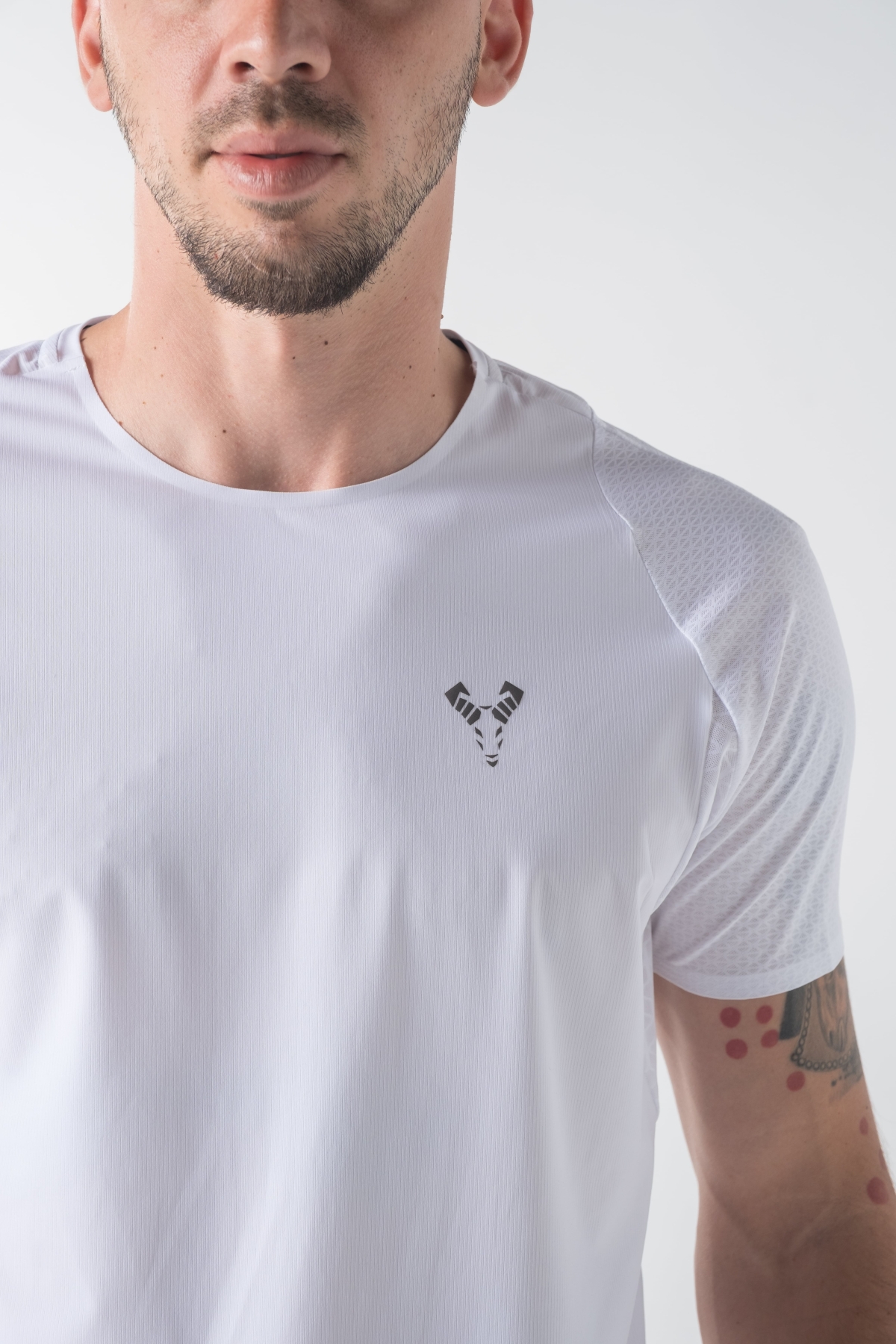 Shift Bluze Meshkujsh – Bluzë sportive me mëngë të shkurtra për meshkuj në ngjyrë të bardhë, me jakë të rrumbullakët, material me teksturë rrjetë për ajrosje, logo minimale Dominer në gjoks, emrin e markës të printuar në pjesën e pasme sipër dhe detaj vertikal me shigjeta në pjesën e poshtme prapa.