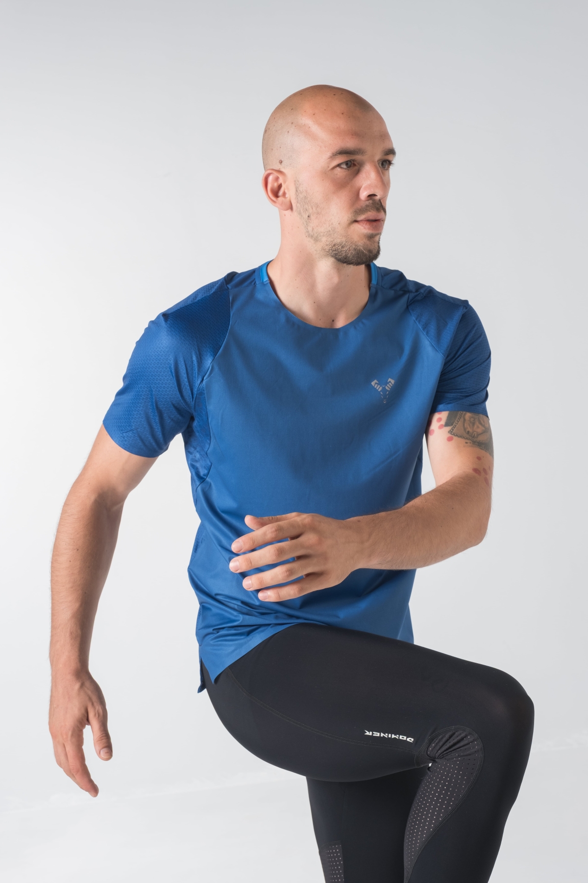 Shift Bluze Meshkujsh – Bluzë sportive me mëngë të shkurtra për meshkuj në ngjyrë të kaltër mbretërore (royal blue), me jakë të rrumbullakët, prerje anësore me të çara të vogla në fund, material me teksturë rrjetë prapa dhe në mëngë për ajrosje, logo minimale Dominer në gjoks, emrin e markës të printuar në pjesën e pasme sipër dhe detaj vertikal me shigjeta në pjesën e poshtme prapa.
