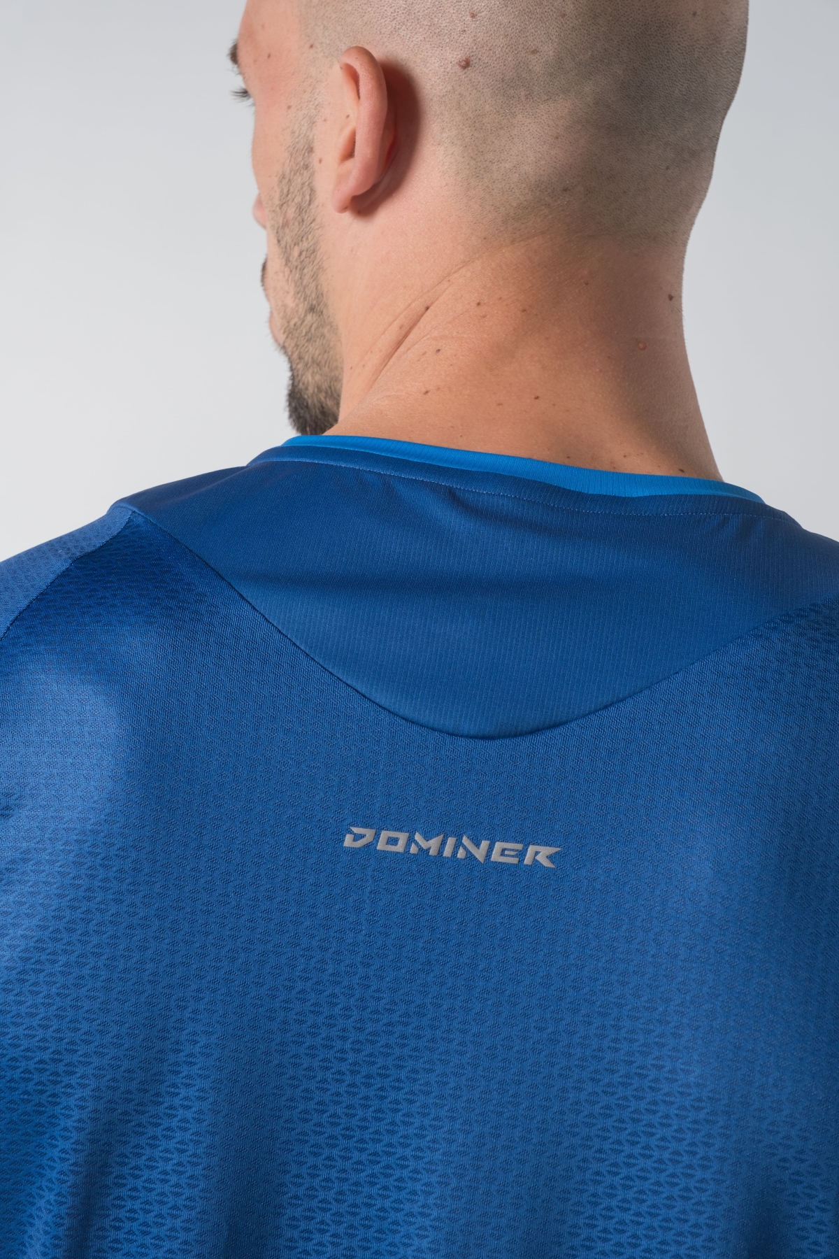 Shift Bluze Meshkujsh – Bluzë sportive me mëngë të shkurtra për meshkuj në ngjyrë të kaltër mbretërore (royal blue), me jakë të rrumbullakët, prerje anësore me të çara të vogla në fund, material me teksturë rrjetë prapa dhe në mëngë për ajrosje, logo minimale Dominer në gjoks, emrin e markës të printuar në pjesën e pasme sipër dhe detaj vertikal me shigjeta në pjesën e poshtme prapa.
