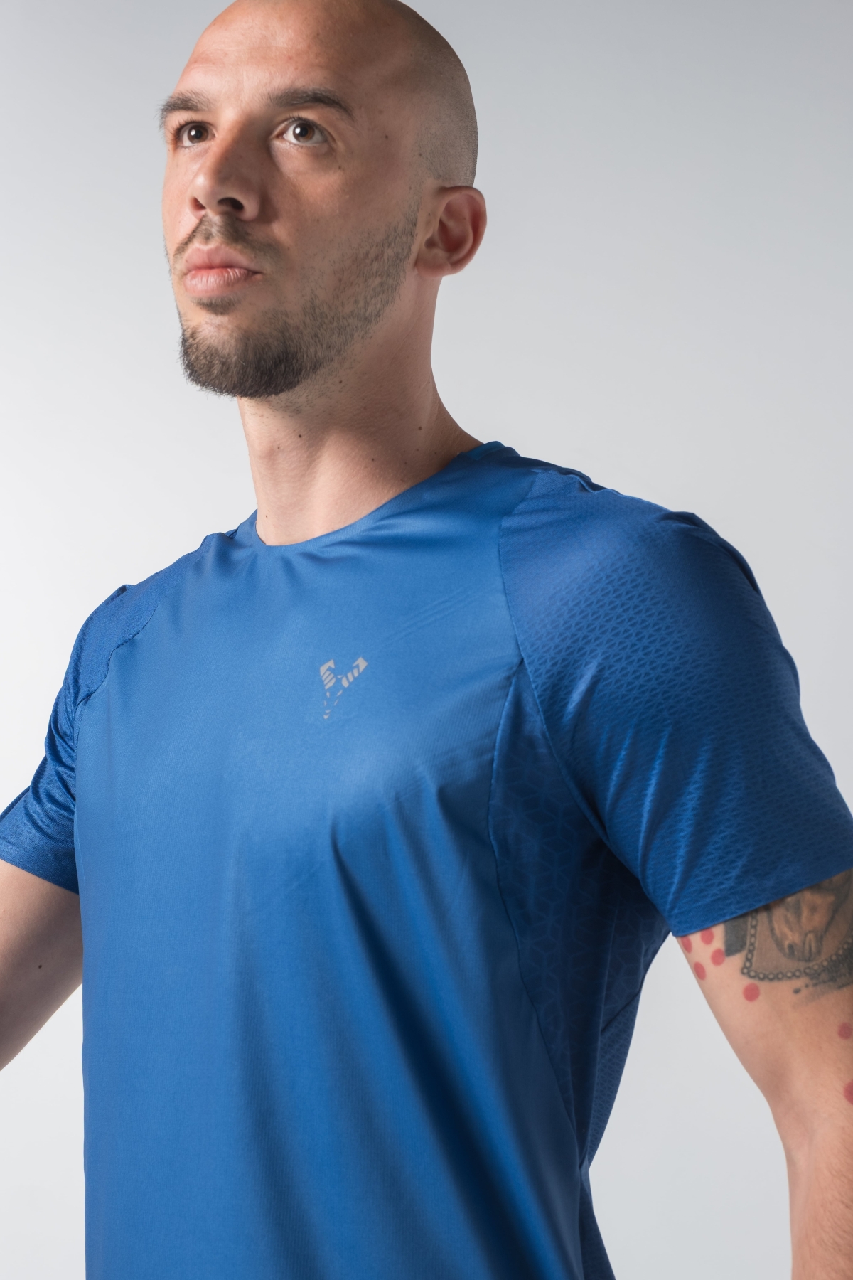 Shift Bluze Meshkujsh – Bluzë sportive me mëngë të shkurtra për meshkuj në ngjyrë të kaltër mbretërore (royal blue), me jakë të rrumbullakët, prerje anësore me të çara të vogla në fund, material me teksturë rrjetë prapa dhe në mëngë për ajrosje, logo minimale Dominer në gjoks, emrin e markës të printuar në pjesën e pasme sipër dhe detaj vertikal me shigjeta në pjesën e poshtme prapa.