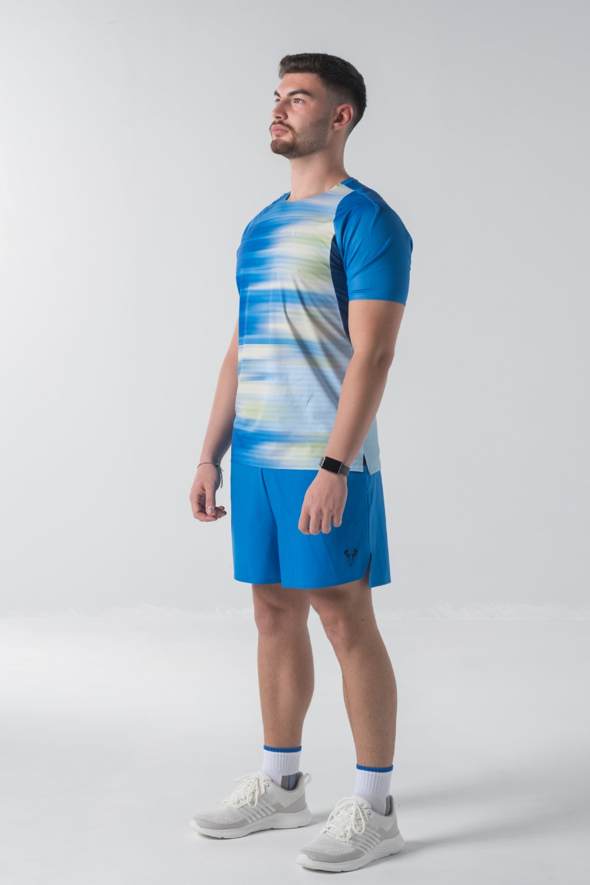 Shift Bluze Meshkujsh – Bluzë sportive me mëngë të shkurtra për meshkuj me print grafik abstrakt me vijëzime horizontale në nuanca të kaltërta, të verdhë e të bardhë, mëngë në ngjyrë të kaltër mbretërore, jakë të rrumbullakët, material me teksturë rrjetë (mesh) me ngjyrë të kaltër të hapur në shpinë për ajrosje, të çara të vogla anësore në fund, logo minimale Dominer në gjoks dhe emrin e markës të printuar në pjesën e pasme sipër.