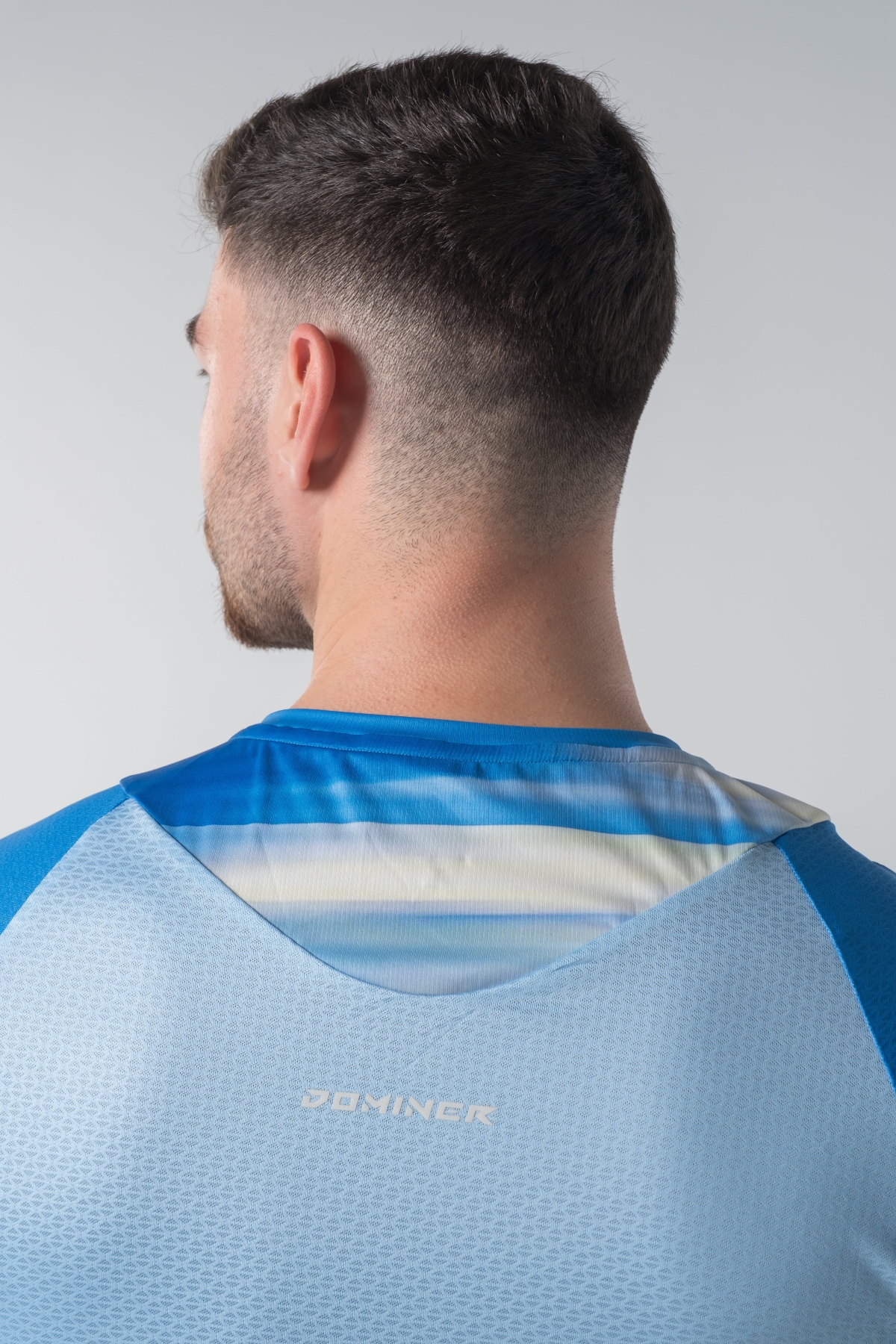 Shift Bluze Meshkujsh – Bluzë sportive me mëngë të shkurtra për meshkuj me print grafik abstrakt me vijëzime horizontale në nuanca të kaltërta, të verdhë e të bardhë, mëngë në ngjyrë të kaltër mbretërore, jakë të rrumbullakët, material me teksturë rrjetë (mesh) me ngjyrë të kaltër të hapur në shpinë për ajrosje, të çara të vogla anësore në fund, logo minimale Dominer në gjoks dhe emrin e markës të printuar në pjesën e pasme sipër.
