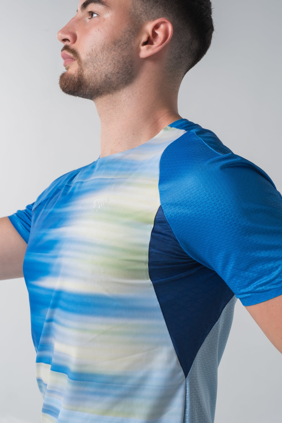 Shift Bluze Meshkujsh – Bluzë sportive me mëngë të shkurtra për meshkuj me print grafik abstrakt me vijëzime horizontale në nuanca të kaltërta, të verdhë e të bardhë, mëngë në ngjyrë të kaltër mbretërore, jakë të rrumbullakët, material me teksturë rrjetë (mesh) me ngjyrë të kaltër të hapur në shpinë për ajrosje, të çara të vogla anësore në fund, logo minimale Dominer në gjoks dhe emrin e markës të printuar në pjesën e pasme sipër.