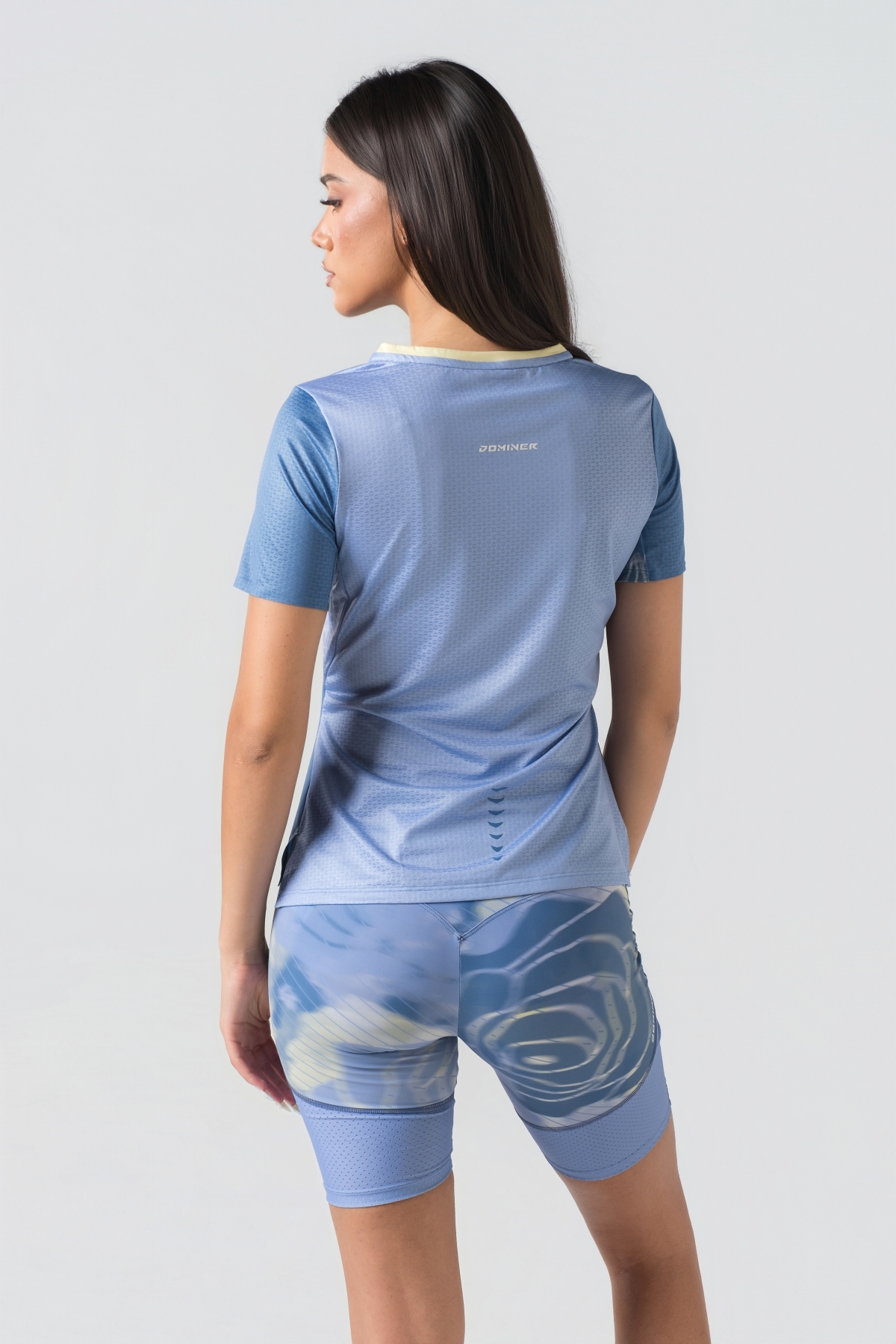 Shift Bluze Femrash – Bluzë sportive me mëngë të shkurtra për femra në ngjyrë të kaltër, me jakë të rrumbullakët me detaj të verdhë pastel, material me teksturë rrjetë (mesh) në shpinë, mëngë dhe panela anësore për ajrosje, të çara të vogla anësore në fund, logo minimale Dominer në gjoks, emrin e markës të printuar në pjesën e pasme sipër dhe detaj vertikal me shigjeta në pjesën e poshtme prapa.
