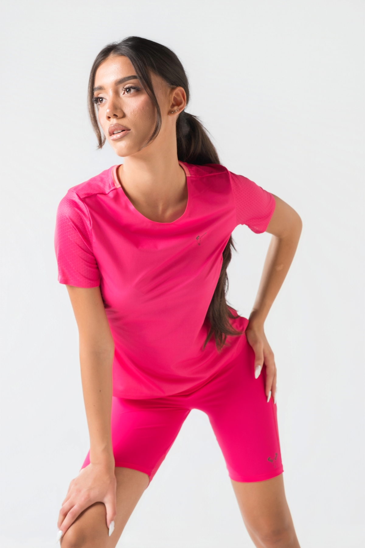 Shift Bluze Femrash – Bluzë sportive me mëngë të shkurtra për femra në ngjyrë rozë elektrike (pink/fuchsia), me jakë të rrumbullakët me detaj rozë më të çelët, material me teksturë rrjetë (mesh) në shpinë, mëngë dhe panela anësore për ajrosje, të çara të vogla anësore në fund, logo minimale Dominer në gjoks dhe emrin e markës të printuar në pjesën e pasme sipër.