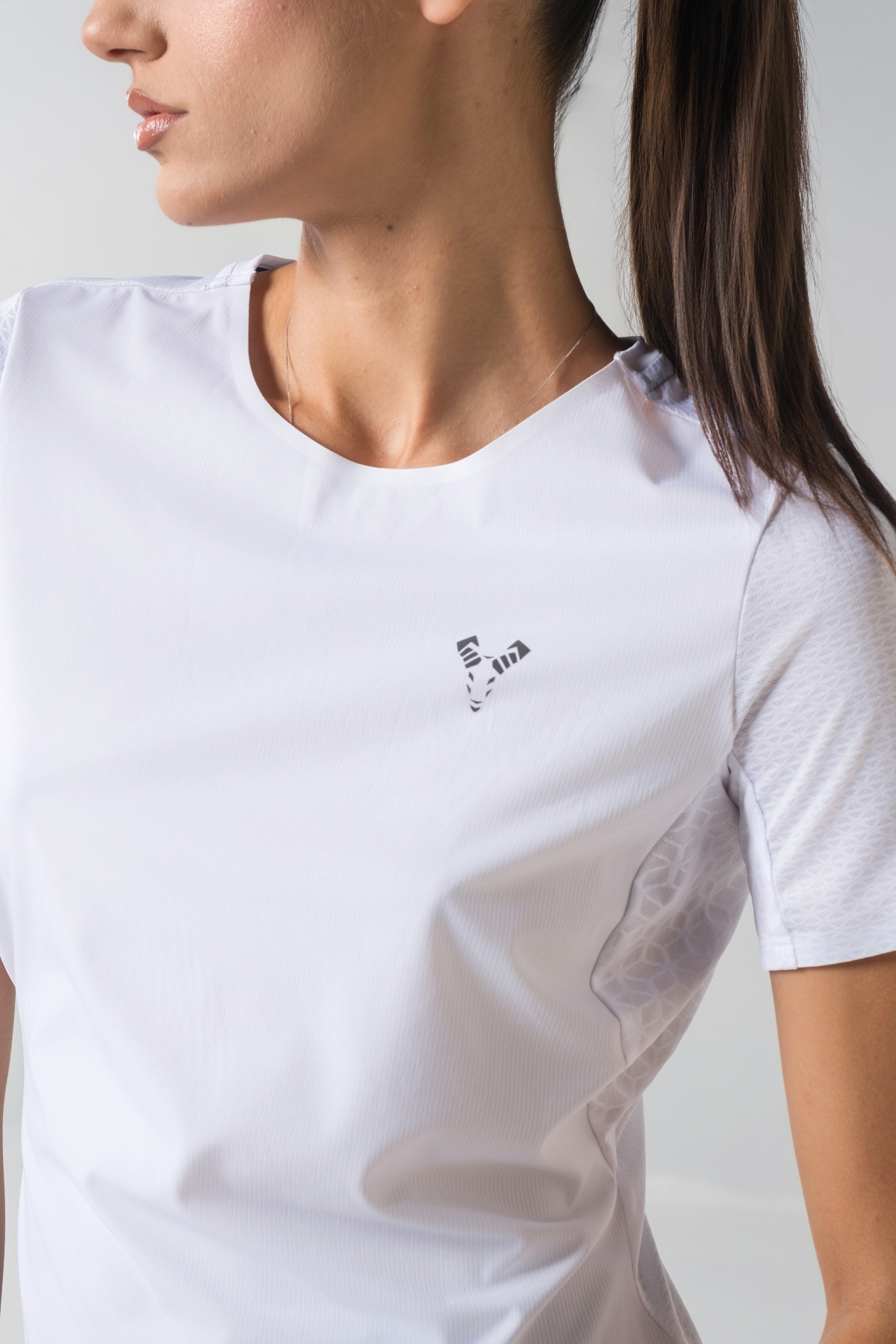 Shift Bluze Femrash – Bluzë sportive me mëngë të shkurtra për femra në ngjyrë të bardhë, me jakë të rrumbullakët, material me teksturë rrjetë (mesh) në shpinë, mëngë dhe panela anësore për ajrosje, të çara të vogla anësore në fund, logo minimale Dominer në gjoks dhe detaj vertikal me shigjeta në pjesën e poshtme prapa.