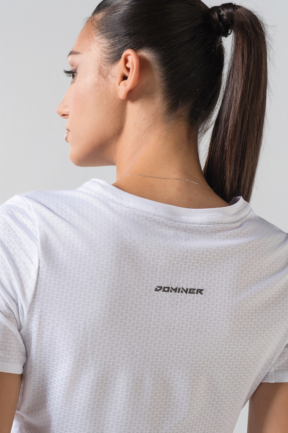 Shift Bluze Femrash – Bluzë sportive me mëngë të shkurtra për femra në ngjyrë të bardhë, me jakë të rrumbullakët, material me teksturë rrjetë (mesh) në shpinë, mëngë dhe panela anësore për ajrosje, të çara të vogla anësore në fund, logo minimale Dominer në gjoks dhe detaj vertikal me shigjeta në pjesën e poshtme prapa.
